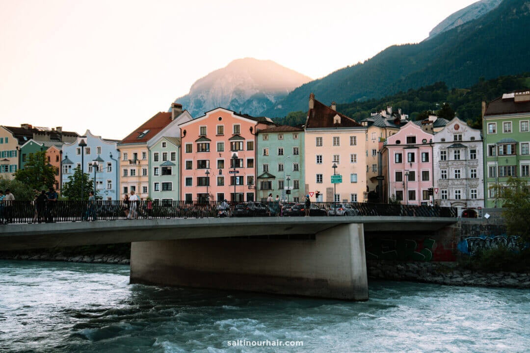 A row of colorful buildings lines the riverbank behind a concrete bridge, with mountains visible in the background at sunsetâ€”one of the many picturesque sights among the things to do in Innsbruck.