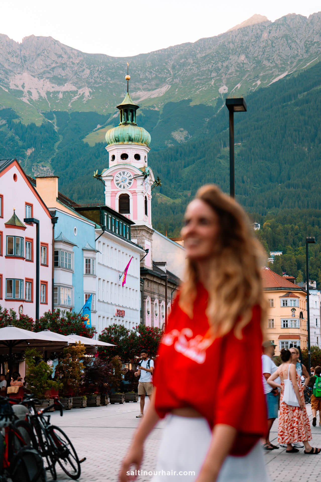 A woman in a red top stands out of focus in the foreground of a European town square in Innsbruck, with colorful buildings and a green-domed towerâ€”capturing the charm and hinting at things to do in Innsbruck against a scenic mountain backdrop.