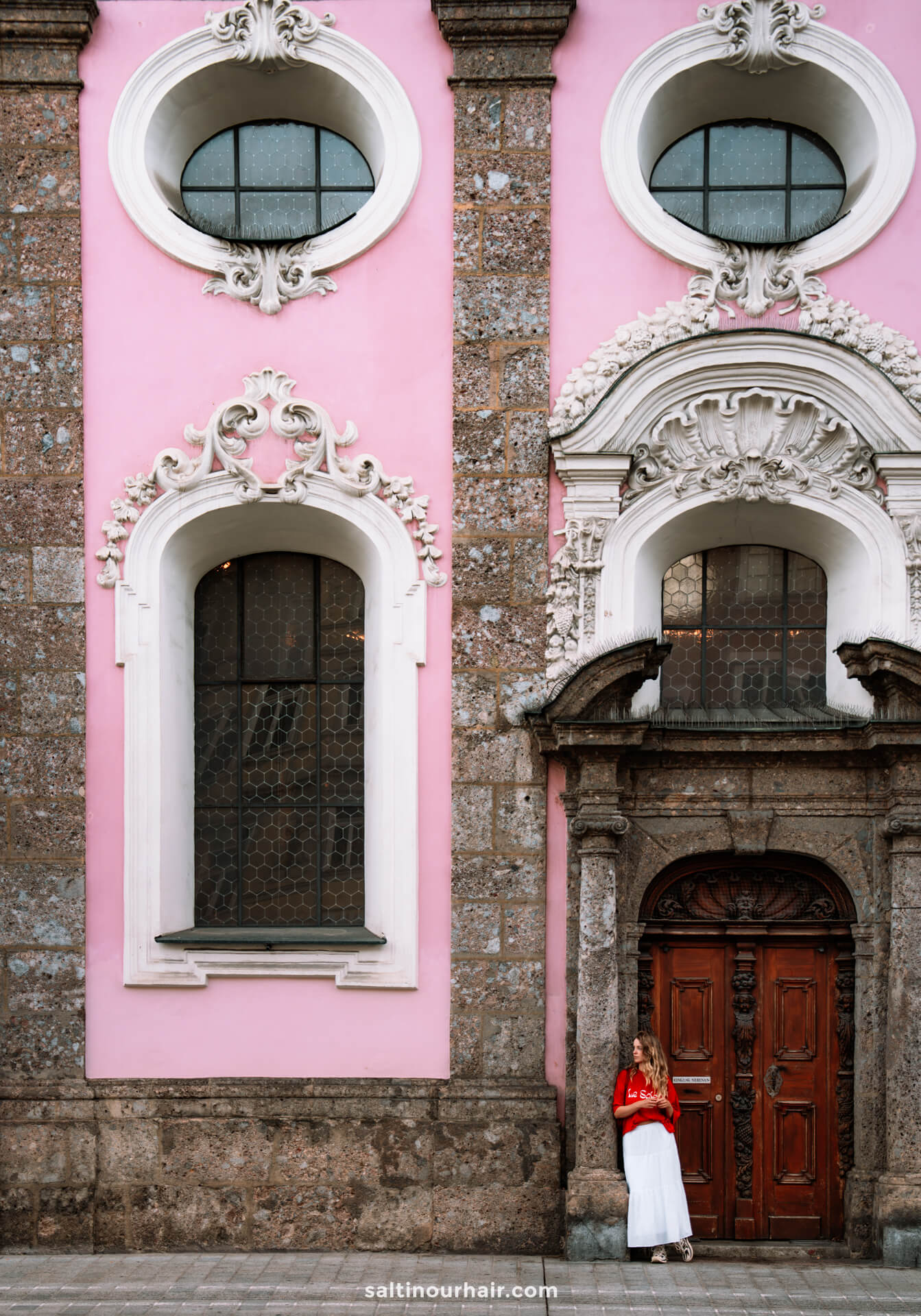 A woman in a red top and white skirt stands by a large wooden door of a pink and brown ornate building with arched windowsâ€”one of the many charming sights to discover among the things to do in Innsbruck, Austria