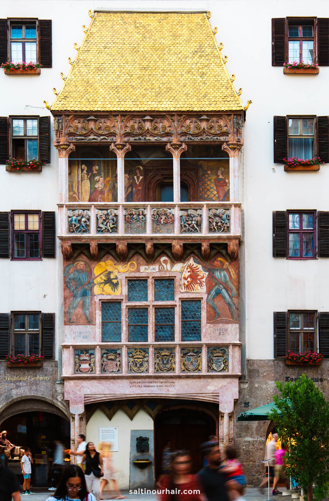 A historic building facade featuring ornate murals, flower boxes on windows, and a prominent golden roofâ€”a must-see among the things to do in Innsbruck; people stroll in front of the entrance.