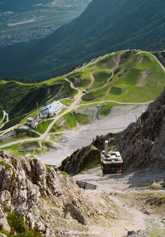A cable car ascends a rocky mountainside, with green hills, winding paths, and buildings visible below in a valleyâ€”one of the unforgettable things to do in Innsbruck.
