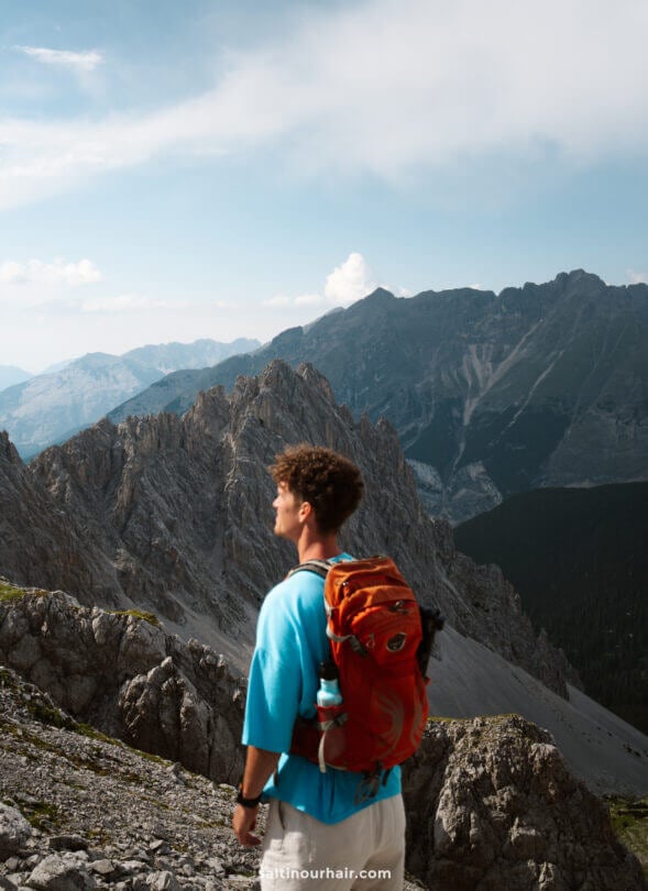 A person with a red backpack stands on a rocky mountain slope, looking at distant peaks under a partly cloudy skyâ€”an inspiring scene reminiscent of the best things to do in Innsbruck for adventurous travelers.