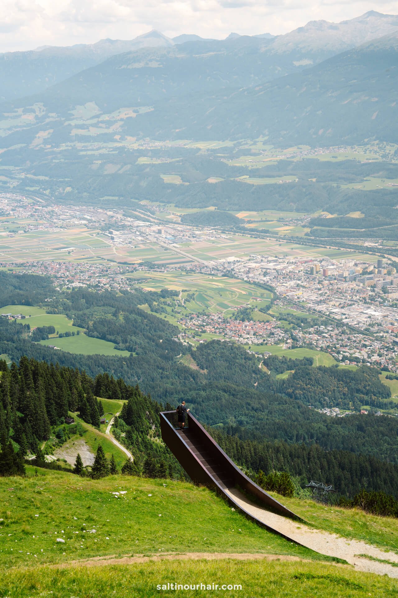 A person stands at the top of a long, metal slide on a grassy hillside overlooking a valley with a town and mountains in the distanceâ€”one of the thrilling things to do in Innsbruck.