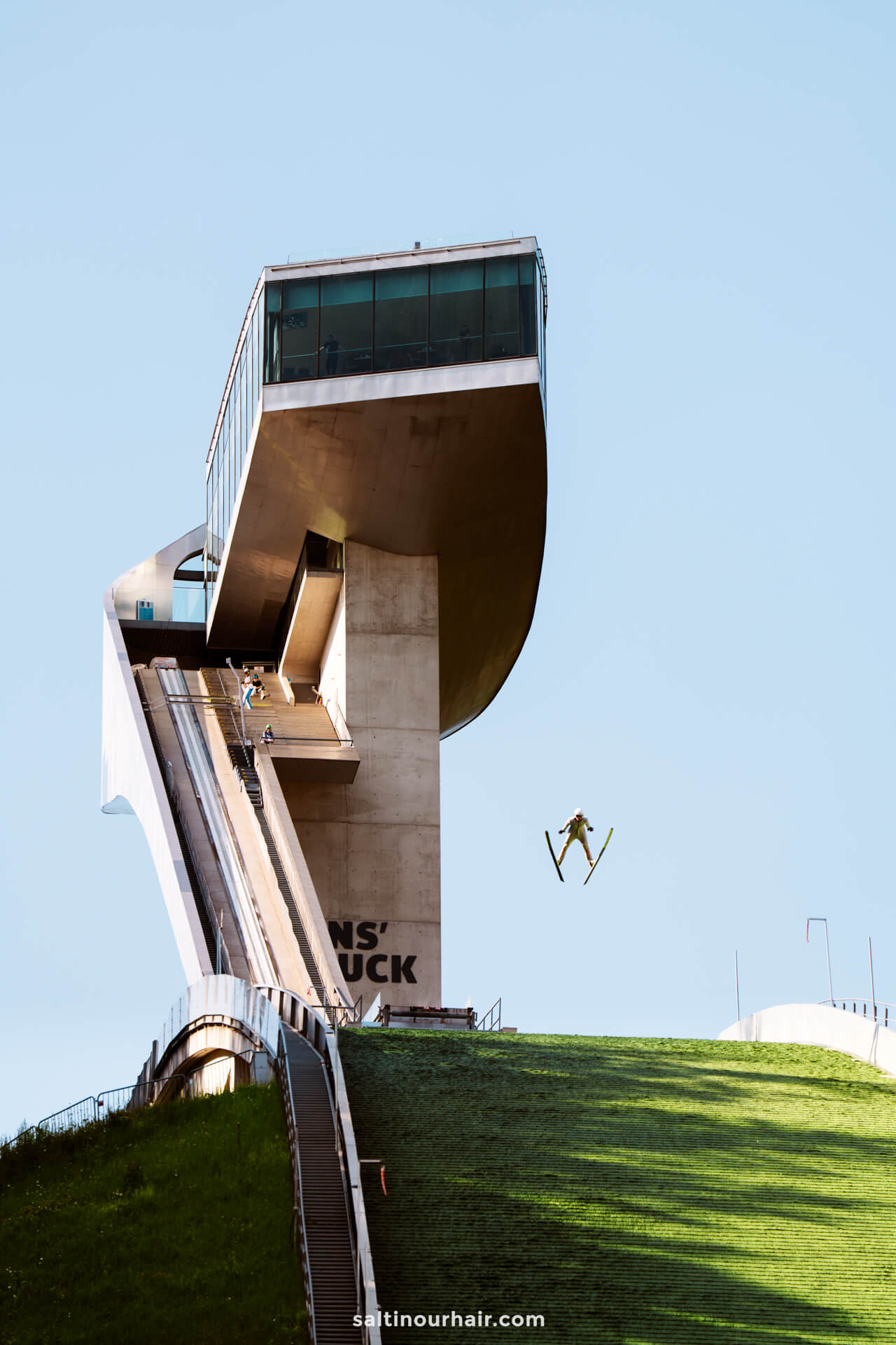 A ski jumper in mid-air descends from a modern ski jumping tower under a clear blue skyâ€”one of the thrilling things to do in Innsbruck, Austria