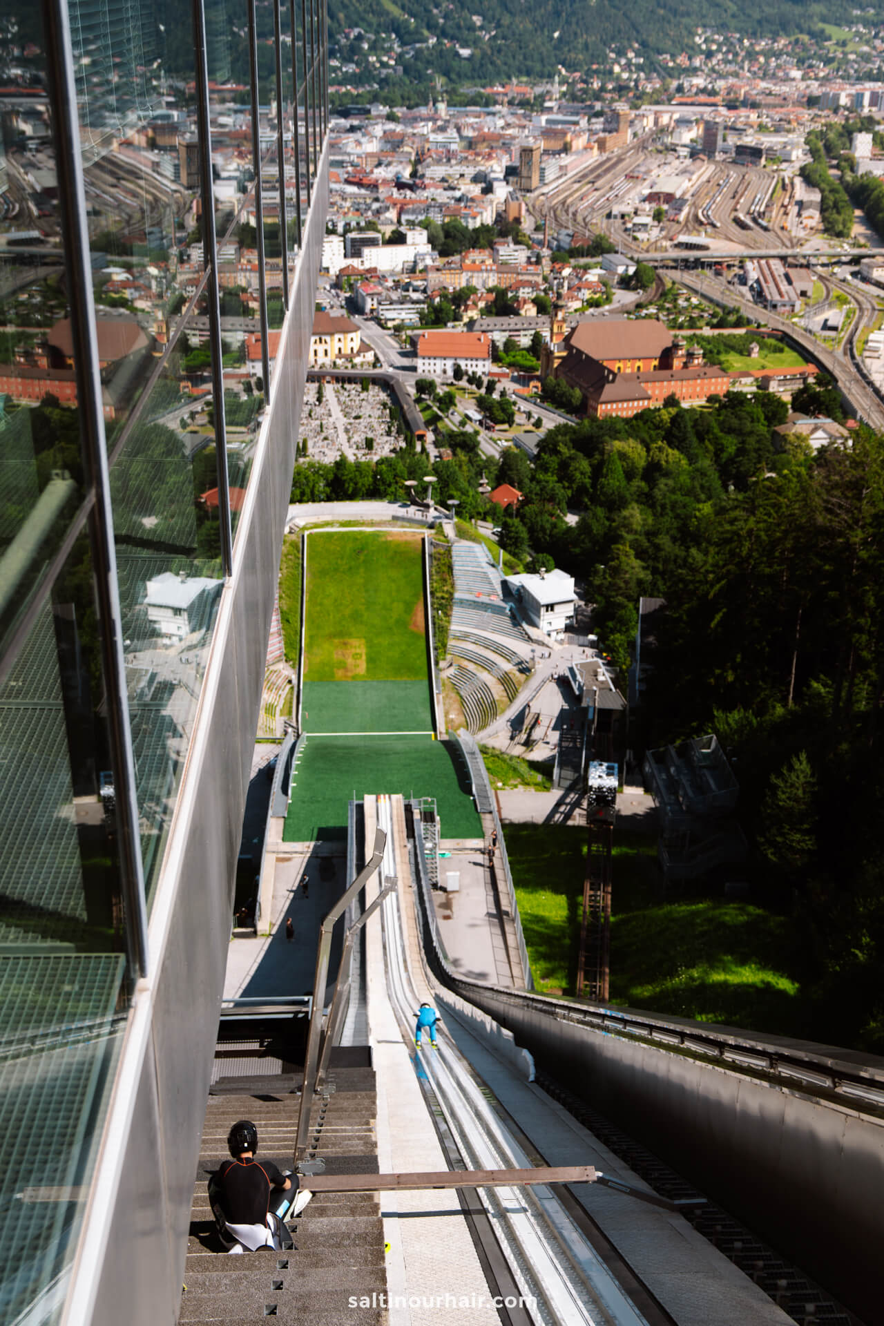 View from the top of a ski jump ramp overlooking a stadium, buildings, and railway tracks, with people on the ramp and a cityscape in the backgroundâ€”one of the must-see things to do in Innsbruck, Austria