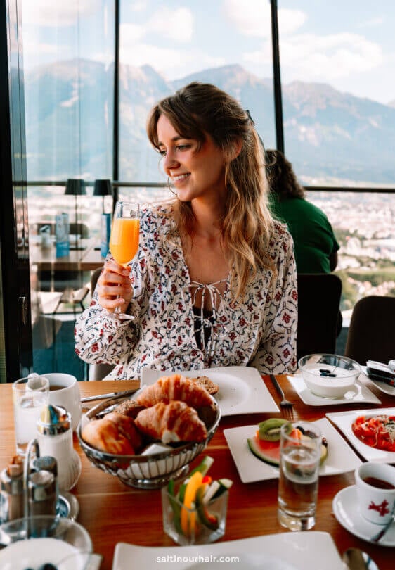 A woman sits at a restaurant table by a window with a mountain view, holding a glass of orange juice. Surrounded by breakfast foods and drinks, she plans her day, exploring things to do in Innsbruck from this scenic spot.