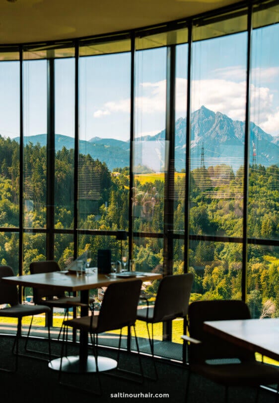 Glass-walled restaurant interior with tables and chairs, overlooking a lush green landscape and distant mountains under a blue skyâ€”an ideal spot to relax after exploring the many things to do in Innsbruck.