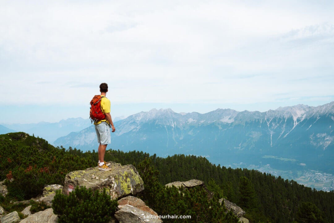 A person wearing a yellow shirt and red backpack stands on a rocky ledge, looking out over a forested valley and distant mountains under a cloudy skyâ€”an inspiring scene for those seeking things to do in Innsbruck.