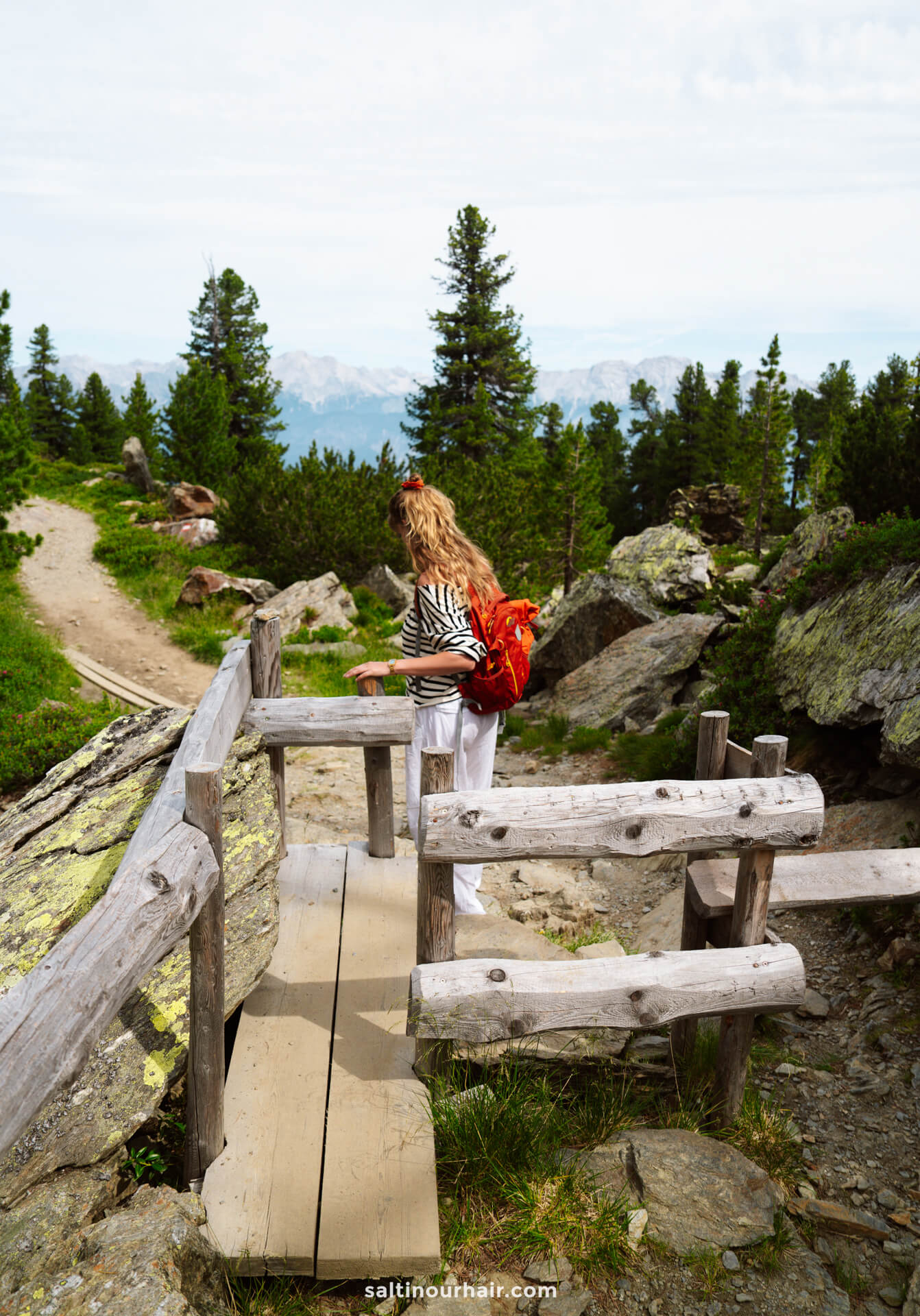 A woman with a red backpack walks across a wooden bridge on a forested mountain trailâ€”one of the scenic things to do in Innsbruckâ€”surrounded by trees and rocks.