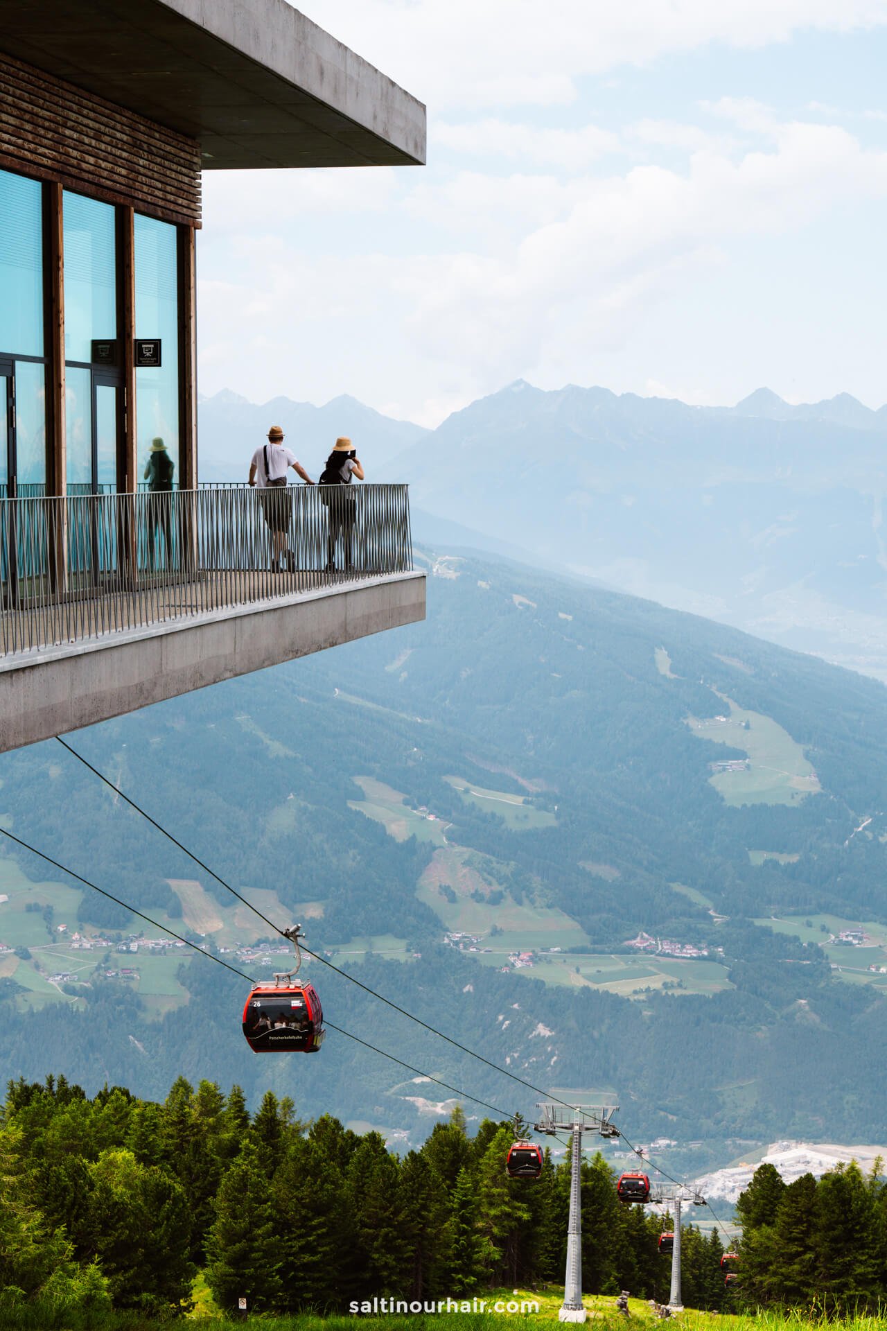 Two people stand on a balcony overlooking a scenic mountain landscape with cable cars belowâ€”one of the top things to do in Innsbruck.