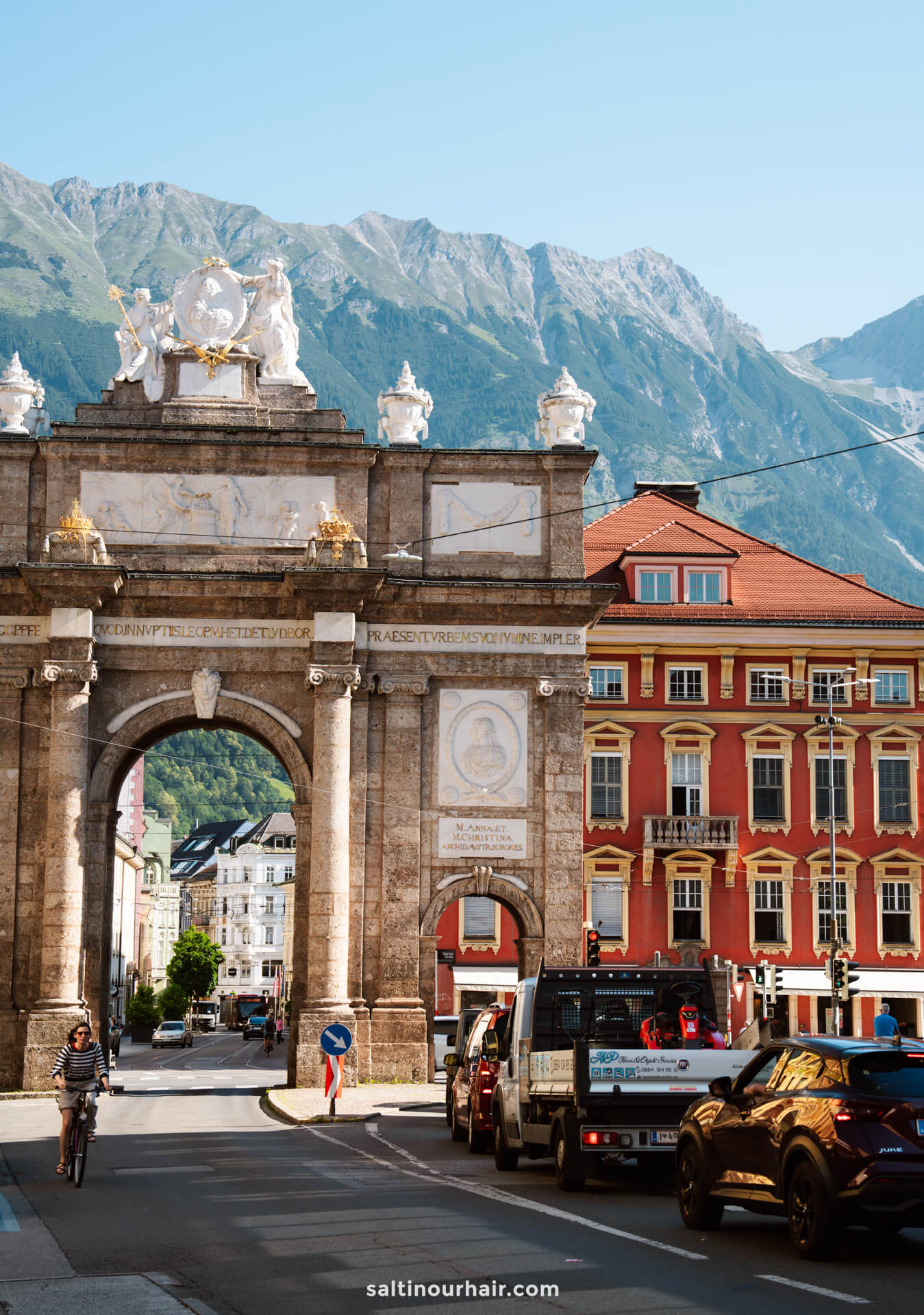 Triumphal arch with ornate statues and columns in a city with colorful buildings, cars, and a cyclist; mountains visible in the backgroundâ€”one of the top things to do in Innsbruck.