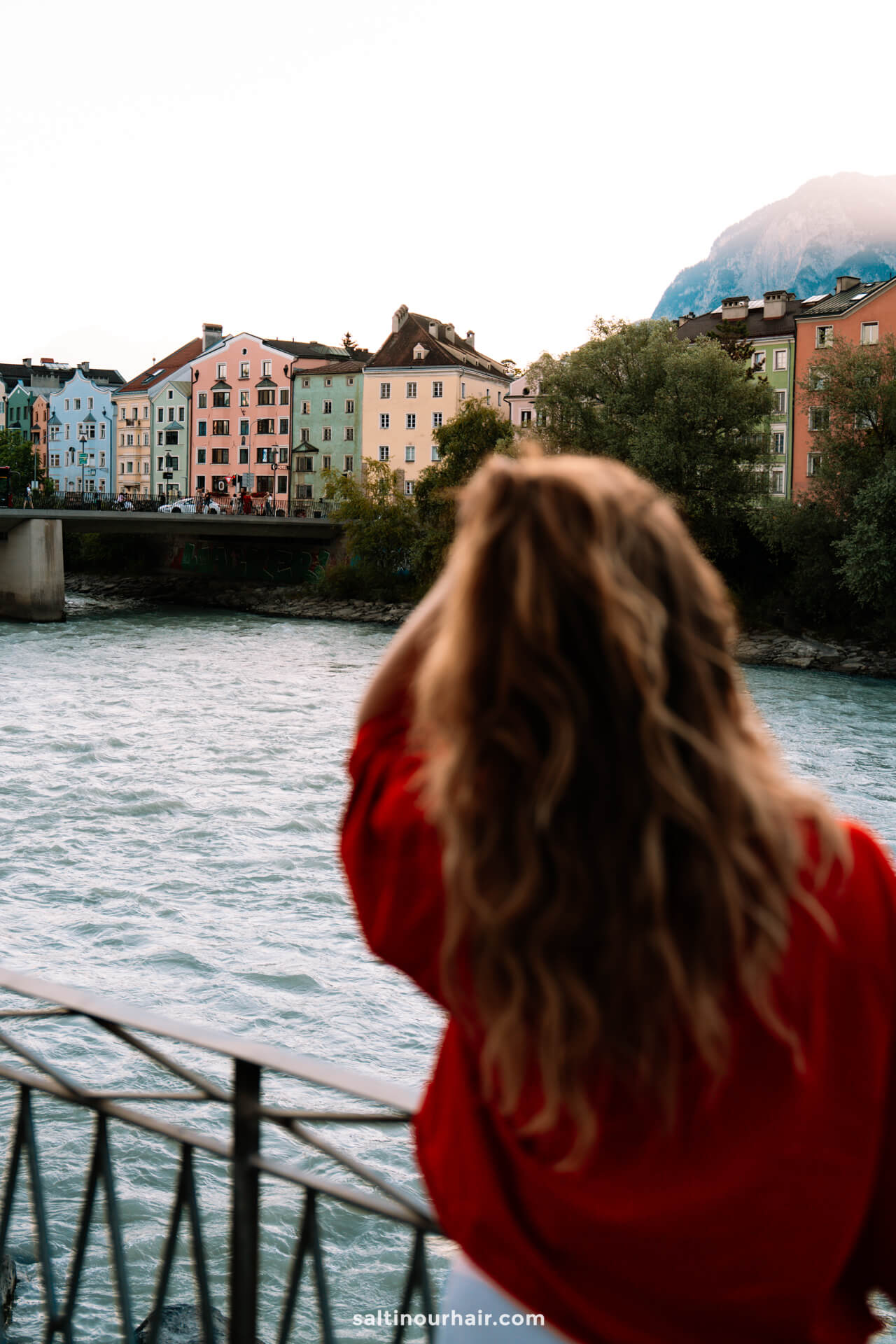 A person with long, wavy hair in a red top stands by a riverside railing in Innsbruck, looking at colorful buildings across the river under a cloudy skyâ€”soaking in one of the classic things to do in Innsbruck.
