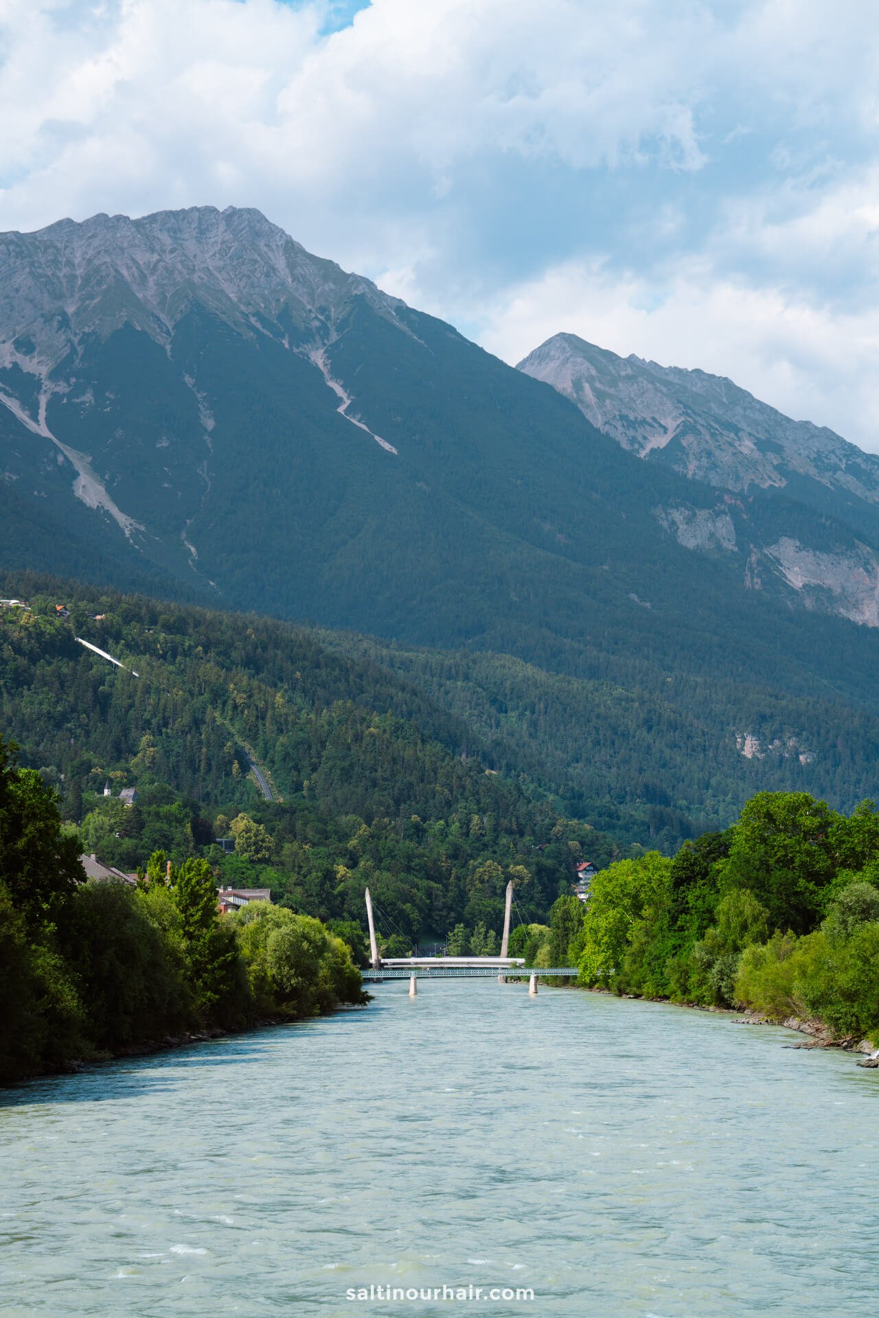 A river flows through a green valley with a bridge in the distance, surrounded by trees and mountains under a partly cloudy skyâ€”perfect for exploring the scenic things to do in Innsbruck.