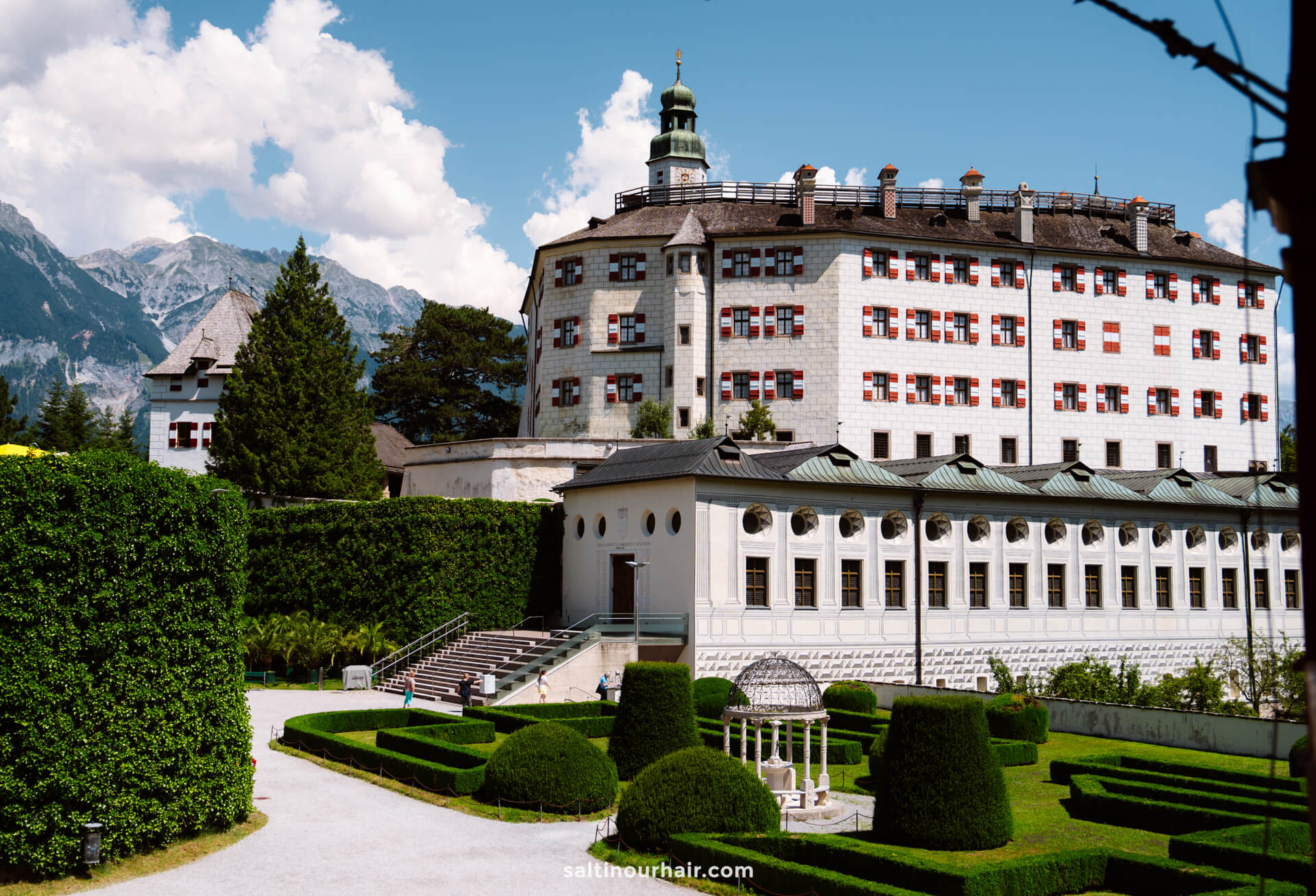 A large, historic building with red-and-white window shutters stands behind formal gardens, trimmed hedges, and a small pavilionâ€”one of the must-see things to do in Innsbruck, with mountains rising in the background.