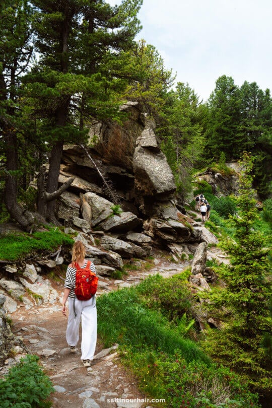 A woman with a red backpack walks along a rocky forest trail, one of the many things to do in Innsbruck, with two more hikers visible ahead among trees and greenery.