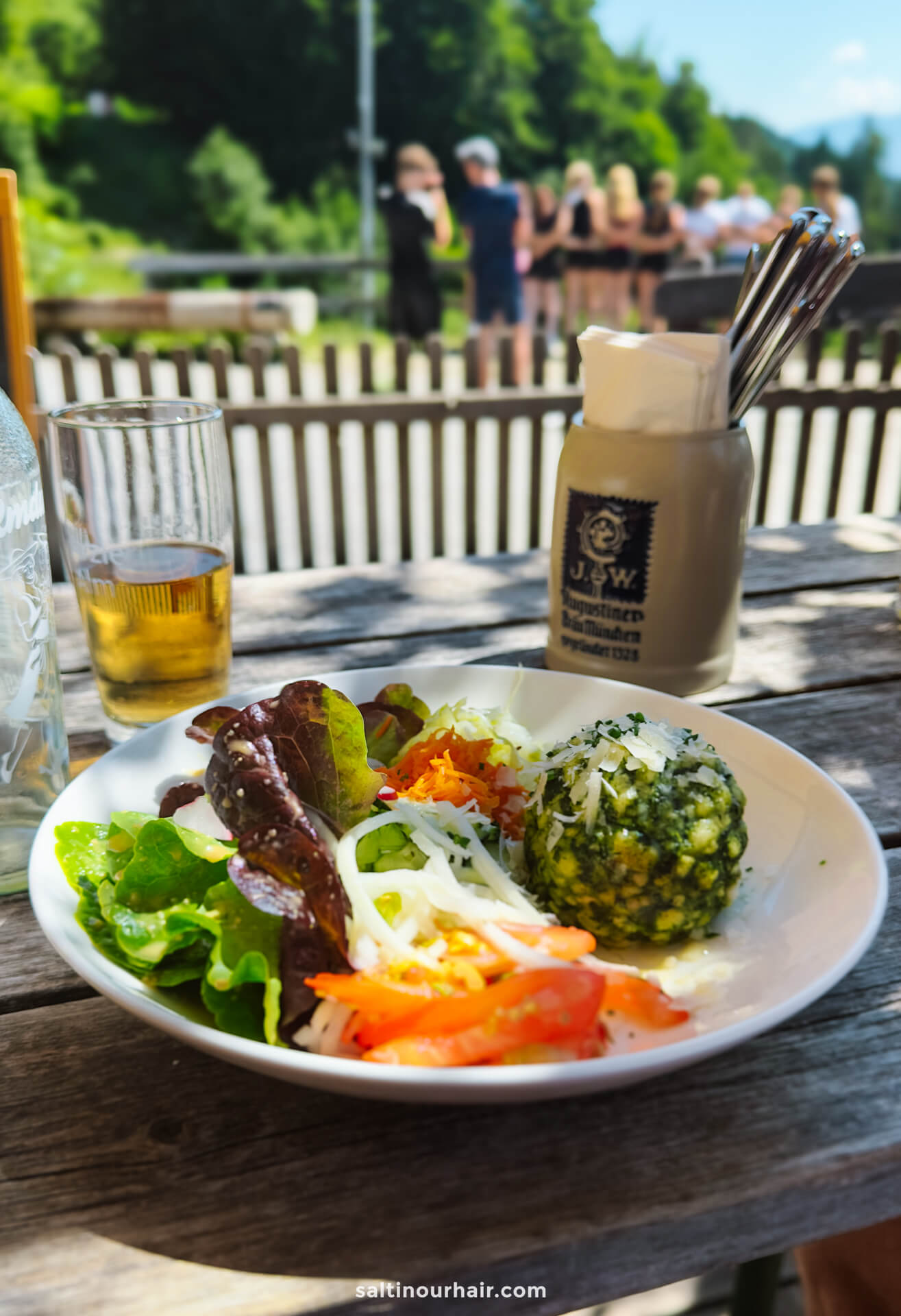 A plate of salad with greens, shredded carrots, cheese, and a round green dumpling sits on an outdoor wooden table beside a drink and utensilsâ€”one of many tasty things to do in Innsbruck, with people and trees in the background.