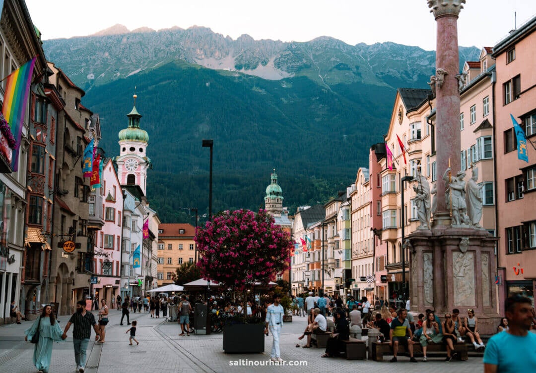A bustling pedestrian street lined with colorful buildings and flags, where people stroll and relax, set against mountain views&mdash;a perfect spot to discover things to do in Innsbruck.