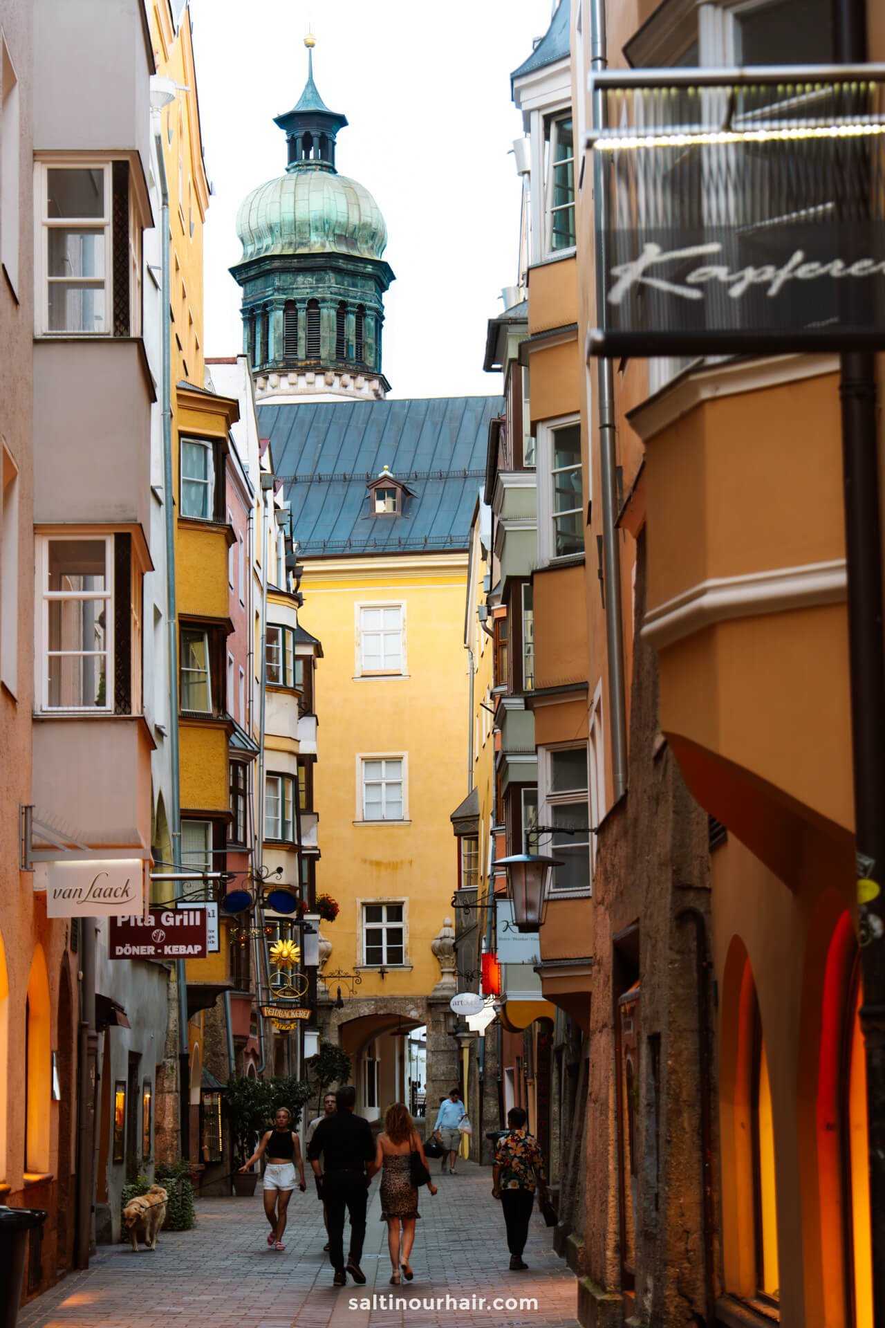 A group of people walk down a narrow street lined with colorful buildings in Innsbruck, with a domed tower visible in the backgroundâ€”one of the many charming things to do in Innsbruck.
