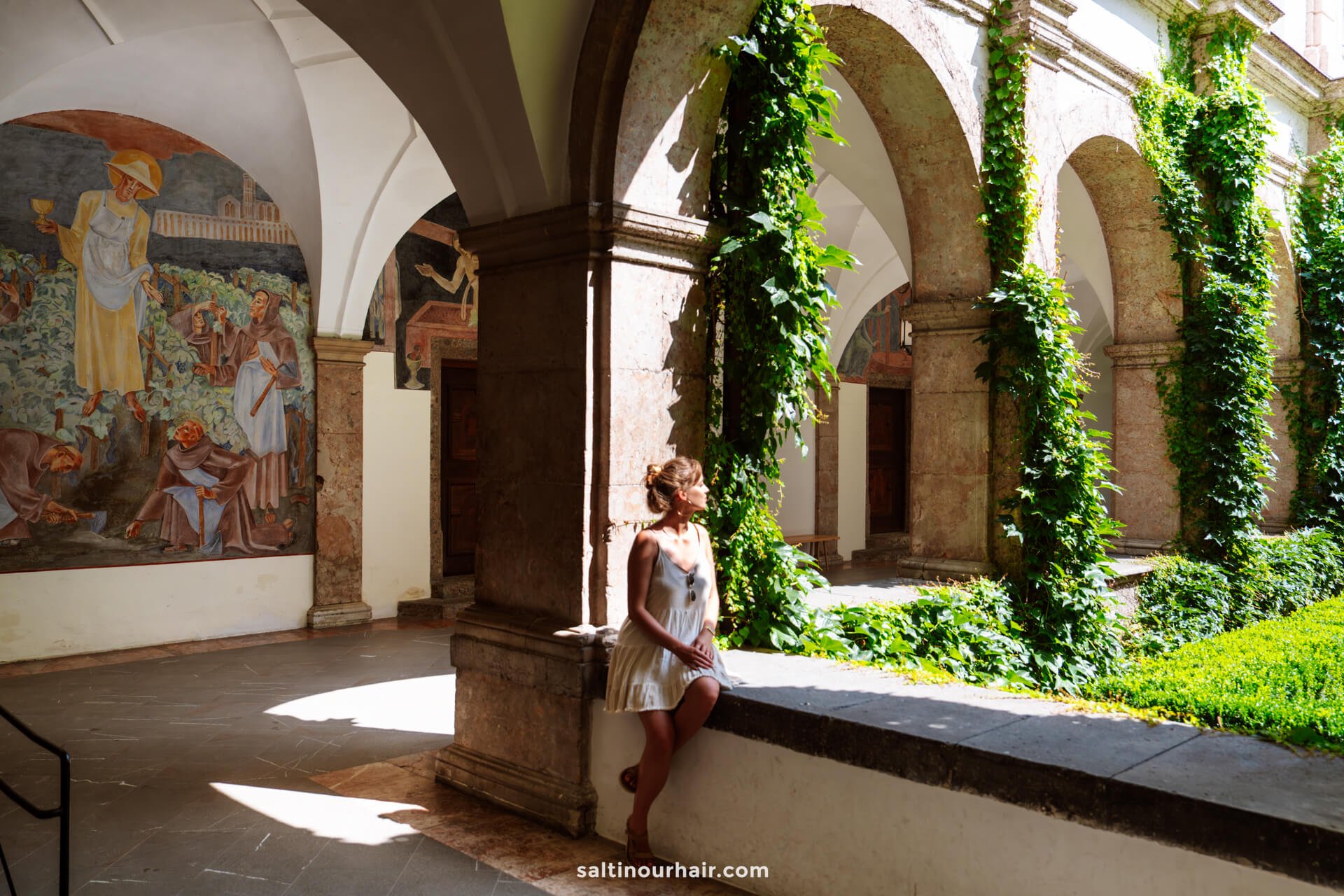 A woman in a white dress sits on a stone ledge in a sunlit cloister with ivy-covered pillars and colorful wall muralsâ€”a serene moment capturing one of the unique things to do in Innsbruck.