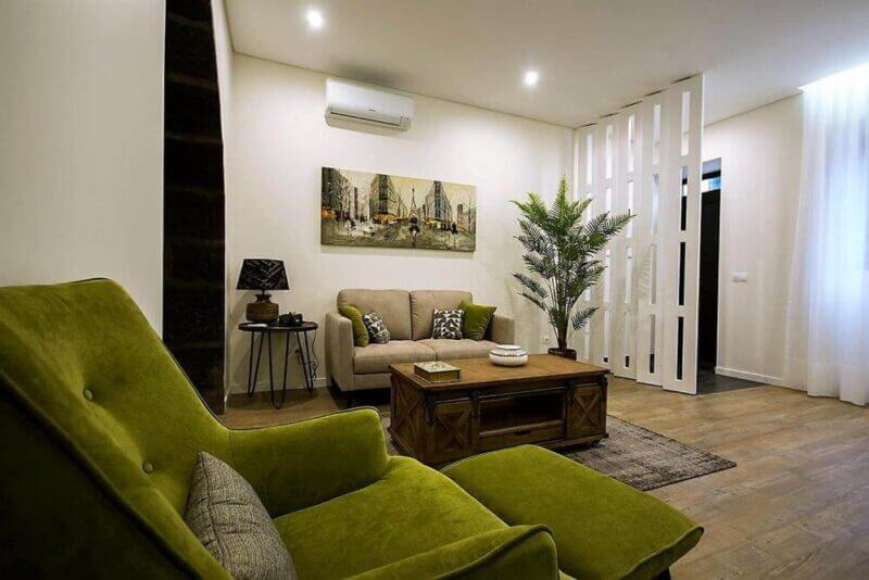 Modern living room with green armchair, beige sofa, wooden coffee table, potted plant from INNature, wall art, decorative lamp, and a room divider under ceiling spotlights—perfect for a Ponta Delgada-inspired retreat.