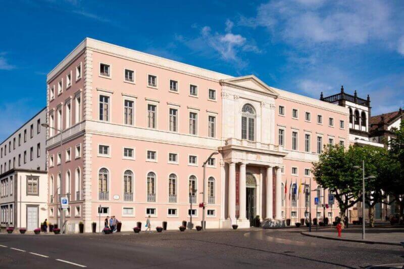 The Vila Gale hotel is a large pink neoclassical building with four columns at the entrance, multiple windows, and flags, situated on a city street in Ponta Delgada under a blue sky.