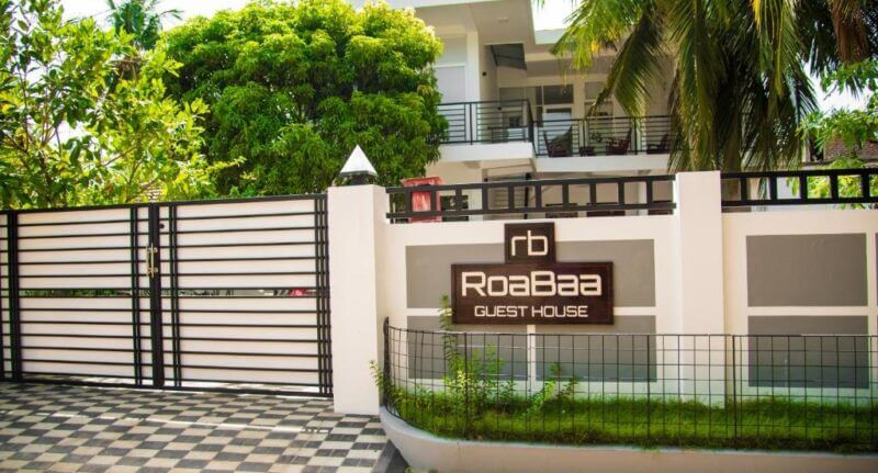 Front view of RoaBaa Guesthouse in Batticaloa, showcasing a modern building, gated entrance, large trees, and a sign displaying the guesthouse name.