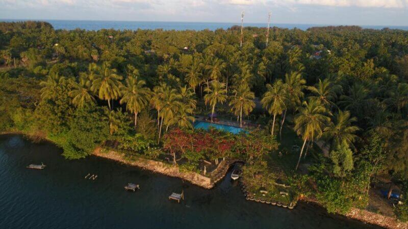 Aerial view of Riviera Resort Batticaloa, featuring a tropical shoreline with dense palm trees, a swimming pool, small docks, and calm water in the foreground.