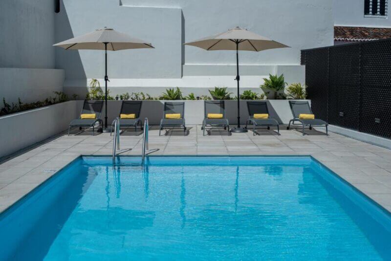 Six lounge chairs with yellow towels and two large umbrellas are arranged beside a clear blue outdoor swimming pool on the tiled deck at Olive Boutique Guesthouse in Sao Miguel.