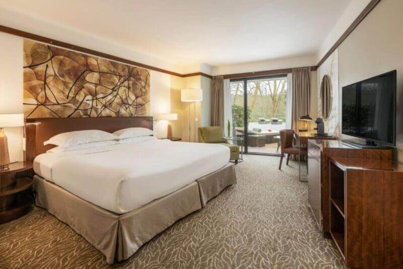 Spacious hotel room at Terra Nostra Garden Hotel on Sao Miguel, featuring a king bed, nightstands, flat-screen TV, armchair, large artwork above the headboard, and a sliding glass door to a patio with serene garden views.