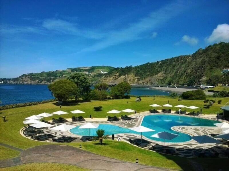 A seaside resort in the Azores, Hotel Azores Pestana Bahia Praia features a large outdoor pool with lounge chairs and white umbrellas, surrounded by green lawns and hills under a blue sky.
