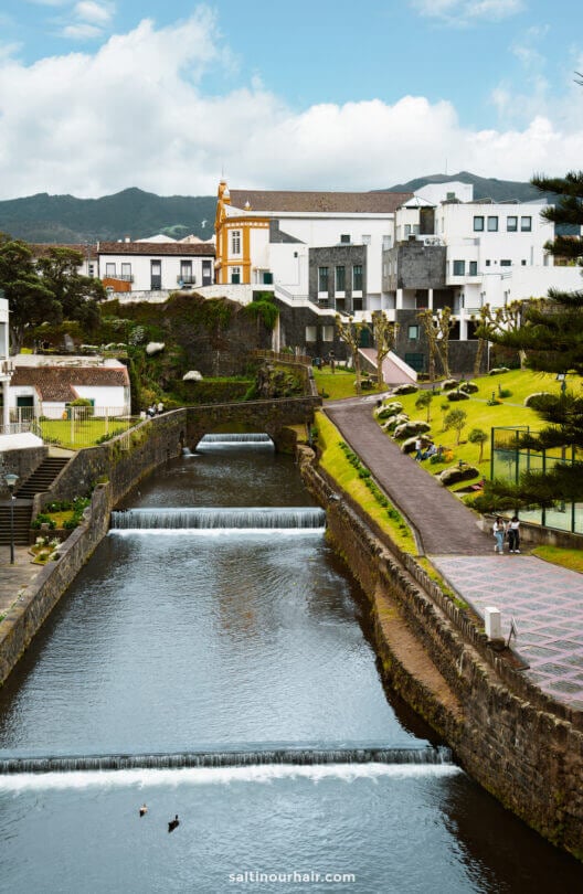 A narrow river with small waterfalls flows through a landscaped town called Ribeira Grande with white buildings, a yellow church, and green hills in the background—an inviting stop on any São Miguel itinerary.