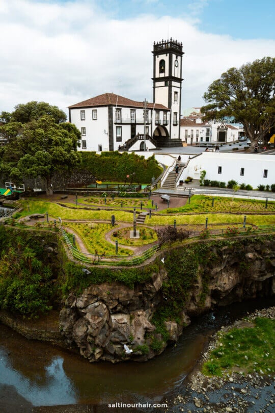 White building with a clock tower in Ribeira Grande beside a lush garden and footbridge, overlooking a rocky stream under a partly cloudy sky—an ideal stop on any São Miguel itinerary.