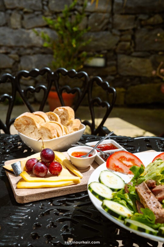 A table set outdoors with sliced bread, grapes, cheese, spreads, and a salad with cucumber, tomato, and lettuce—perfect for a relaxing pause during your São Miguel itinerary. Black metal chair and stone wall in the background.