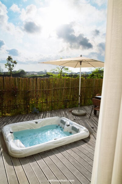 A white outdoor hot tub sits on a wooden deck with bubbling water, next to a sun umbrella, bordered by a bamboo fence—an inviting spot to unwind on your São Miguel itinerary, with scenic sky and greenery in the background.