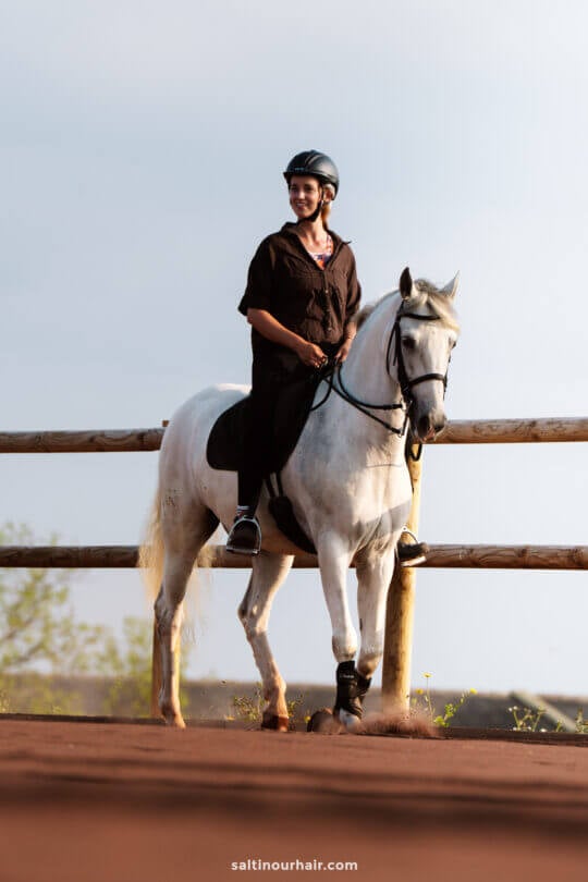 A woman wearing a helmet and dark clothing rides a white horse along a fenced dirt path outdoors at Lusitano Garden Villas, adding an adventurous touch to any São Miguel itinerary.