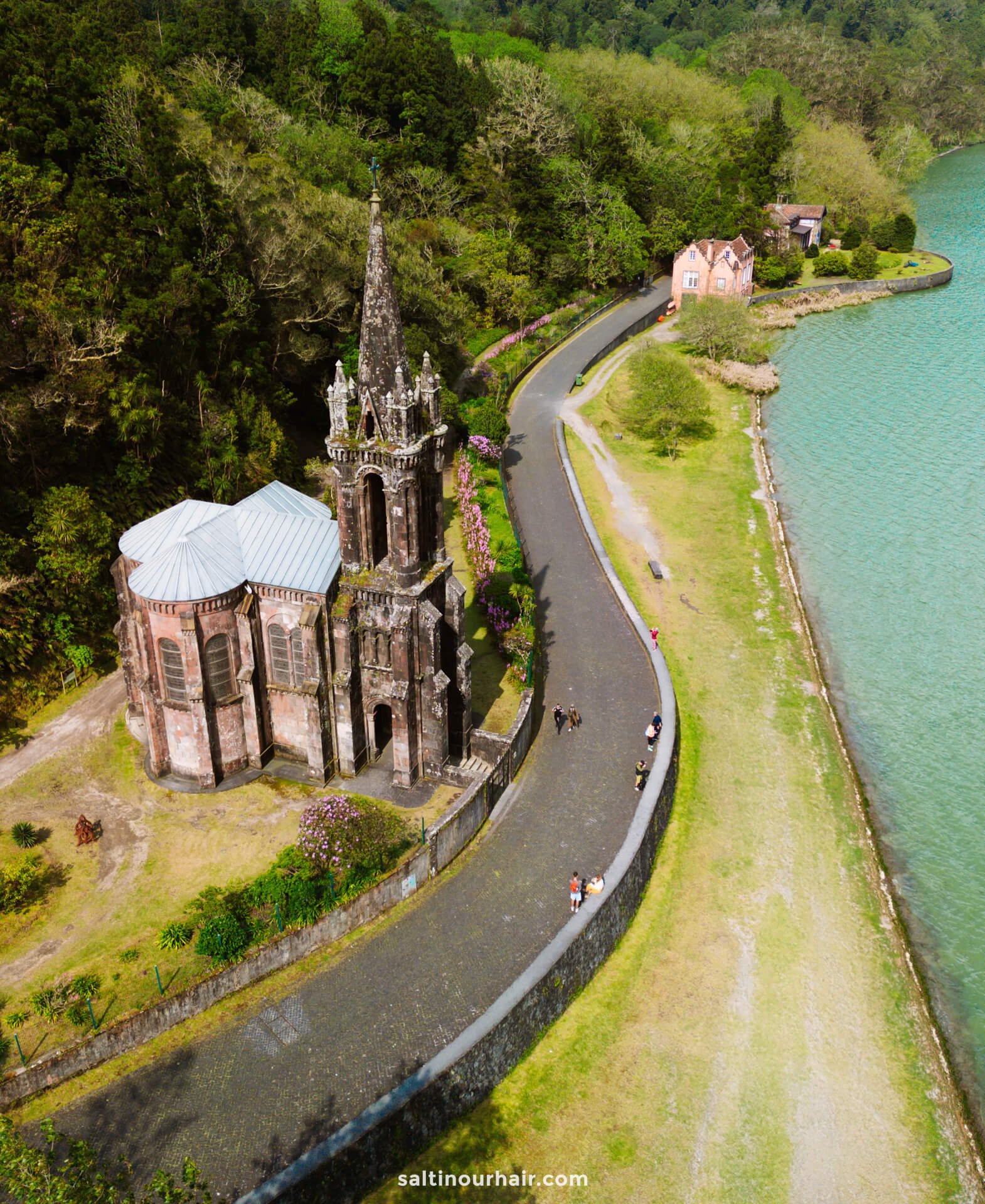 Aerial view of a historic stone church called Capela de Nossa Senhora das Vitórias with a tall spire beside a winding road next to a turquoise lake, surrounded by greenery—a must-see highlight on any São Miguel itinerary.