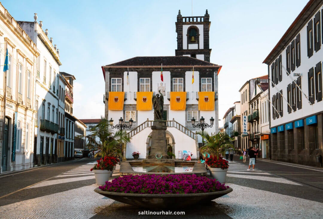 A town square in Ponta Delgada with a round flower bed and fountain in the center, facing a historic building adorned with flags and statues, flanked by white buildings—a lovely stop on any São Miguel azores itinerary.