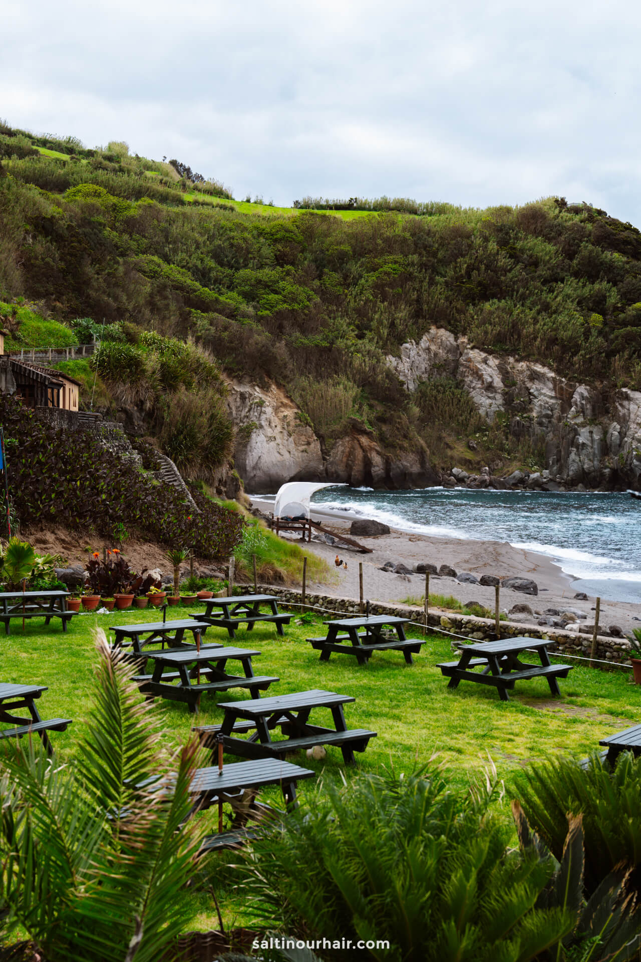 Picnic tables on a grassy area overlook a rocky beach and ocean, with green hills and cliffs in the background - Praia dos Moinhos is one of the best beaches in sao miguel azores