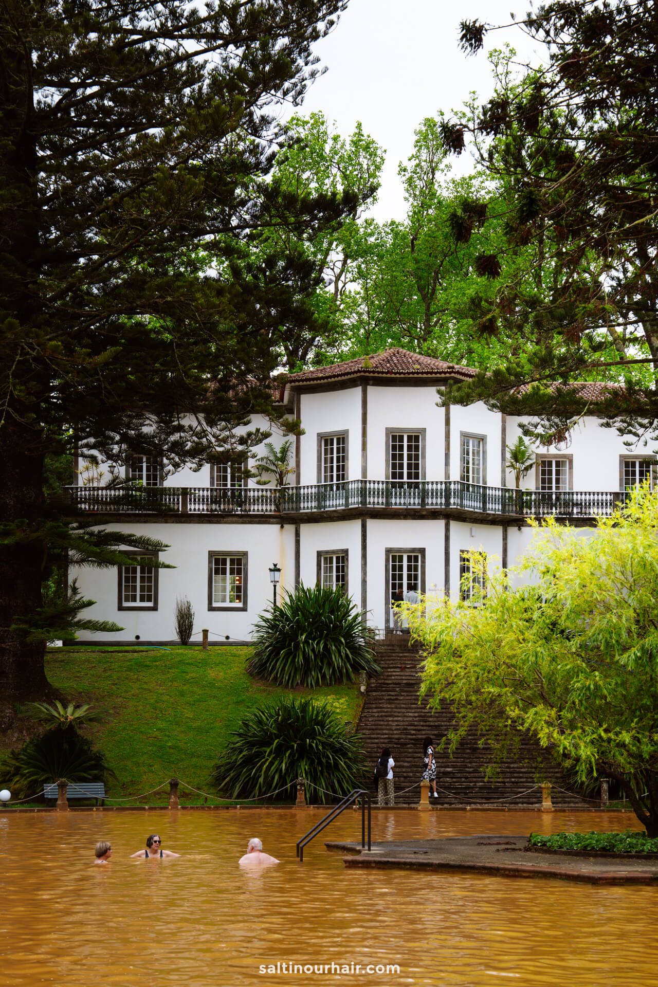A white colonial-style building with large windows stands behind trees and a brown-colored outdoor thermal pool where several people are swimming—one of the must-see things to do in Furnas.