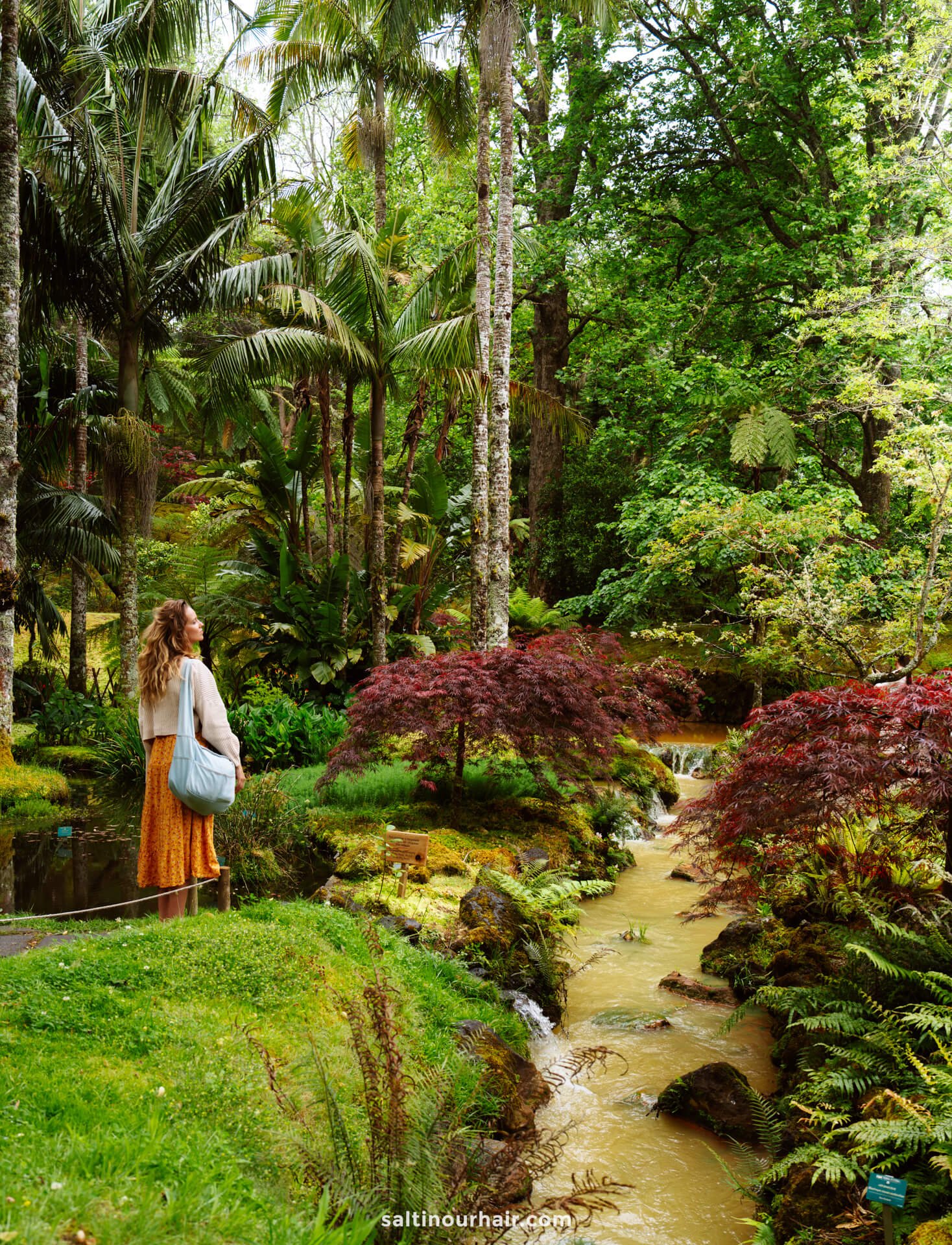A woman with a backpack and an orange skirt stands on a lush, green path beside a stream surrounded by tropical plants—one of the many scenic things to do in Furnas.