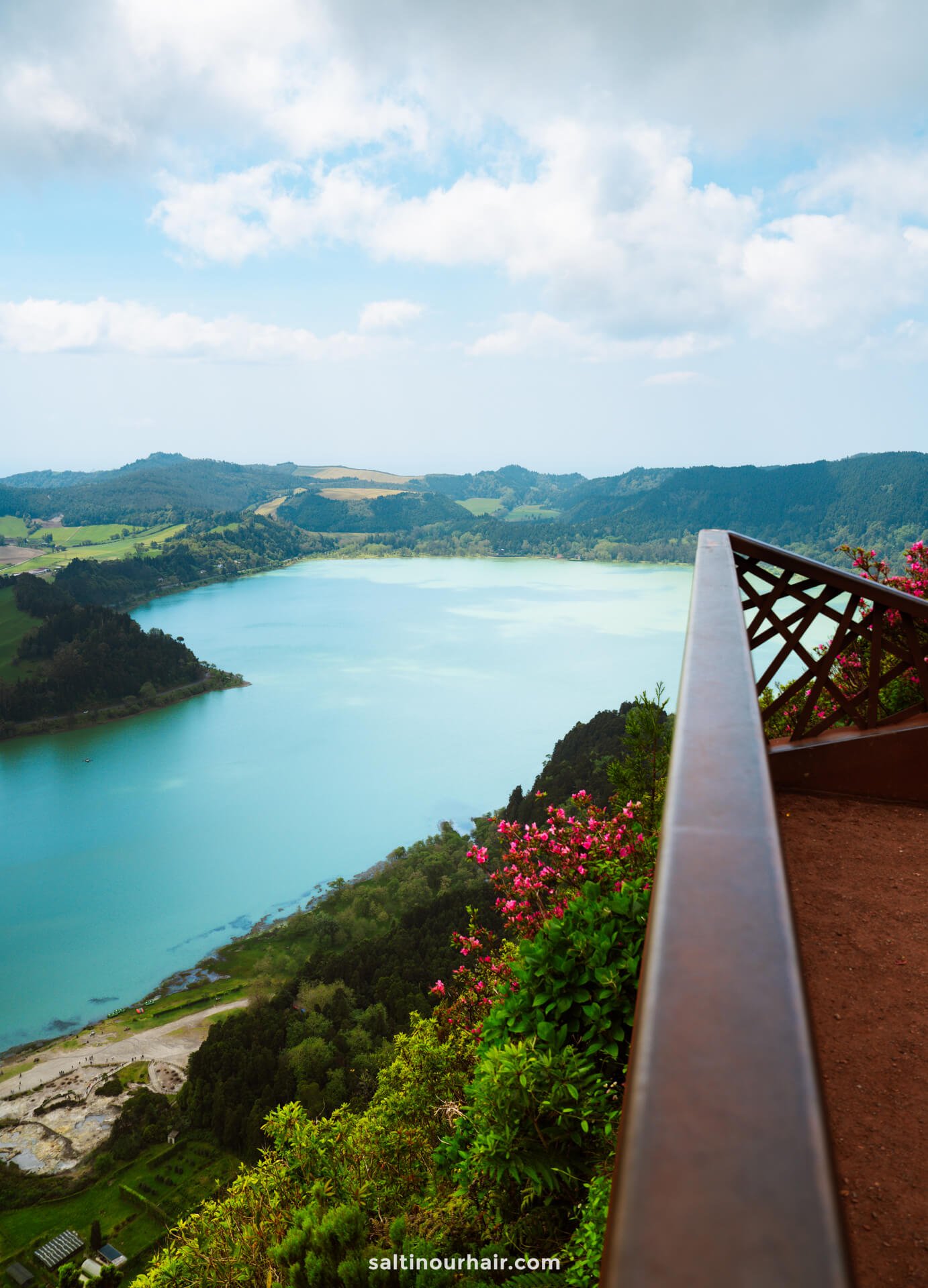 A scenic view of a turquoise lake surrounded by lush green hills, seen from a wooden lookout with flowers in the foreground—a must-see among things to do in Furnas.