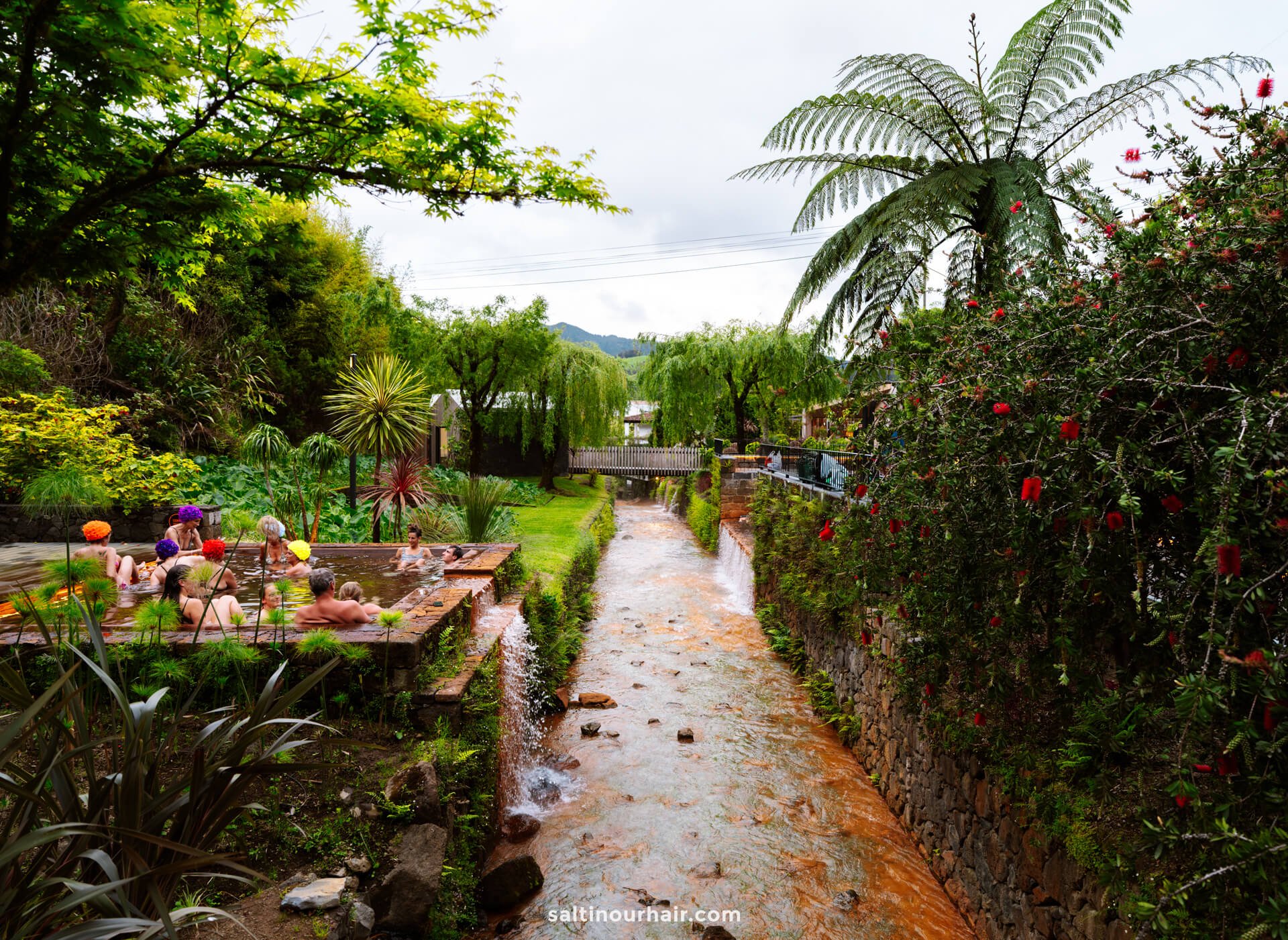 People are relaxing in outdoor hot spring pools beside a small, orange-colored stream, surrounded by lush green foliage and flowering plants—one of the top things to do in Furnas.