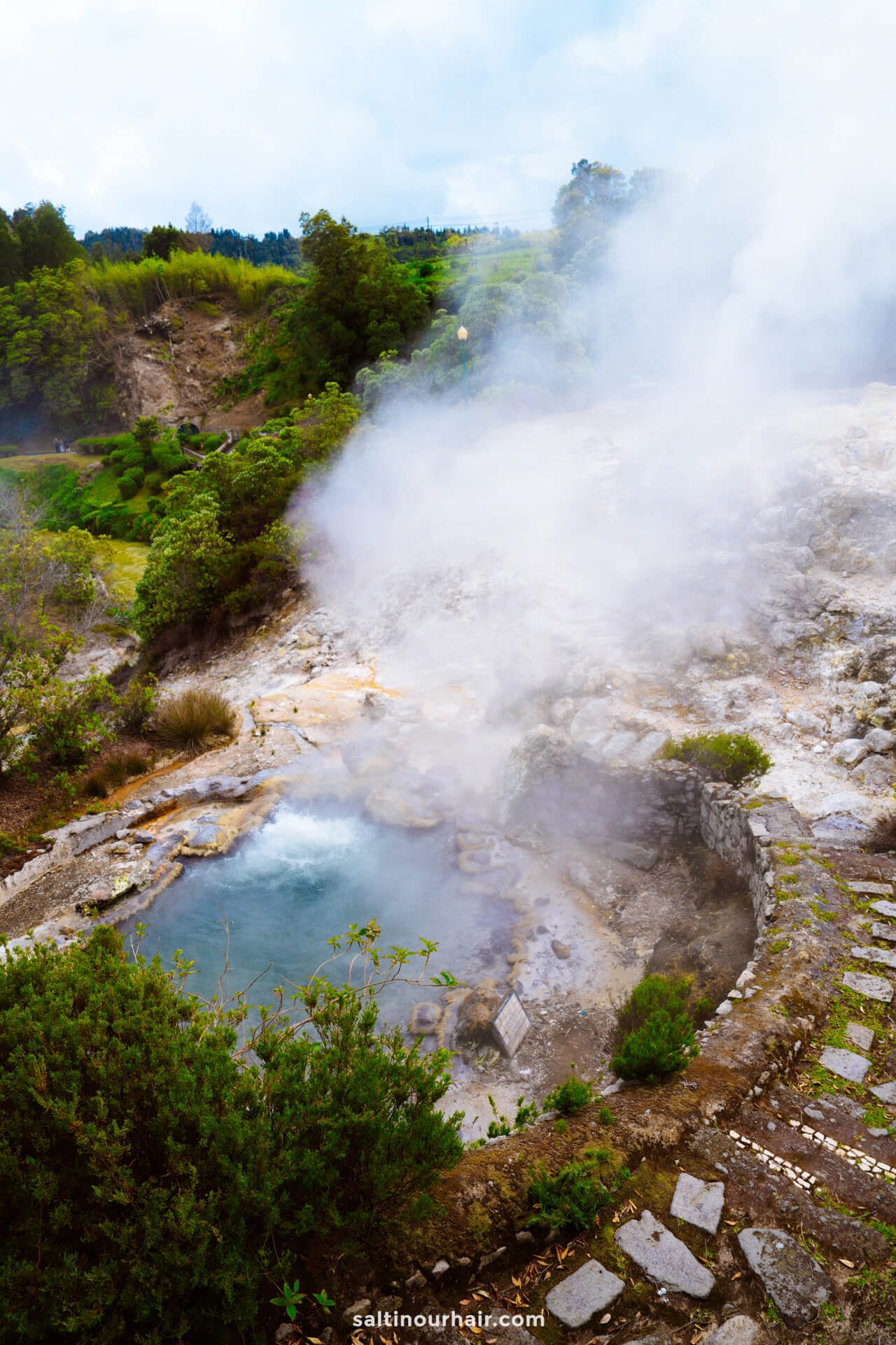 A steaming hot spring pool surrounded by rocks and greenery, with stone steps leading down and thick steam rising into the air—one of the unforgettable things to do in Furnas.