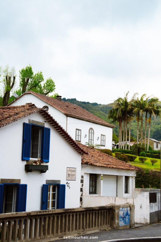 White house with blue shutters and a tiled roof nestled in front of a green Furnas hillside with palm trees and another white building in the background.
