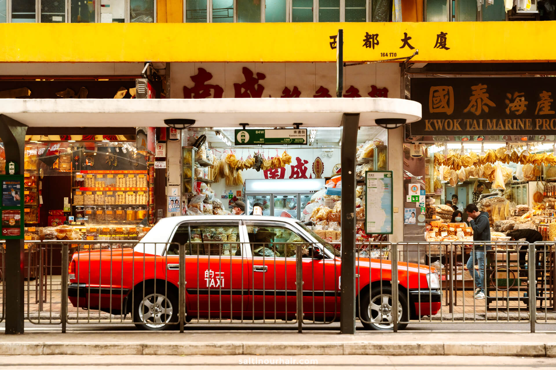 A red taxi is parked in front of shops selling dried goods and seafood on a busy Hong Kong street