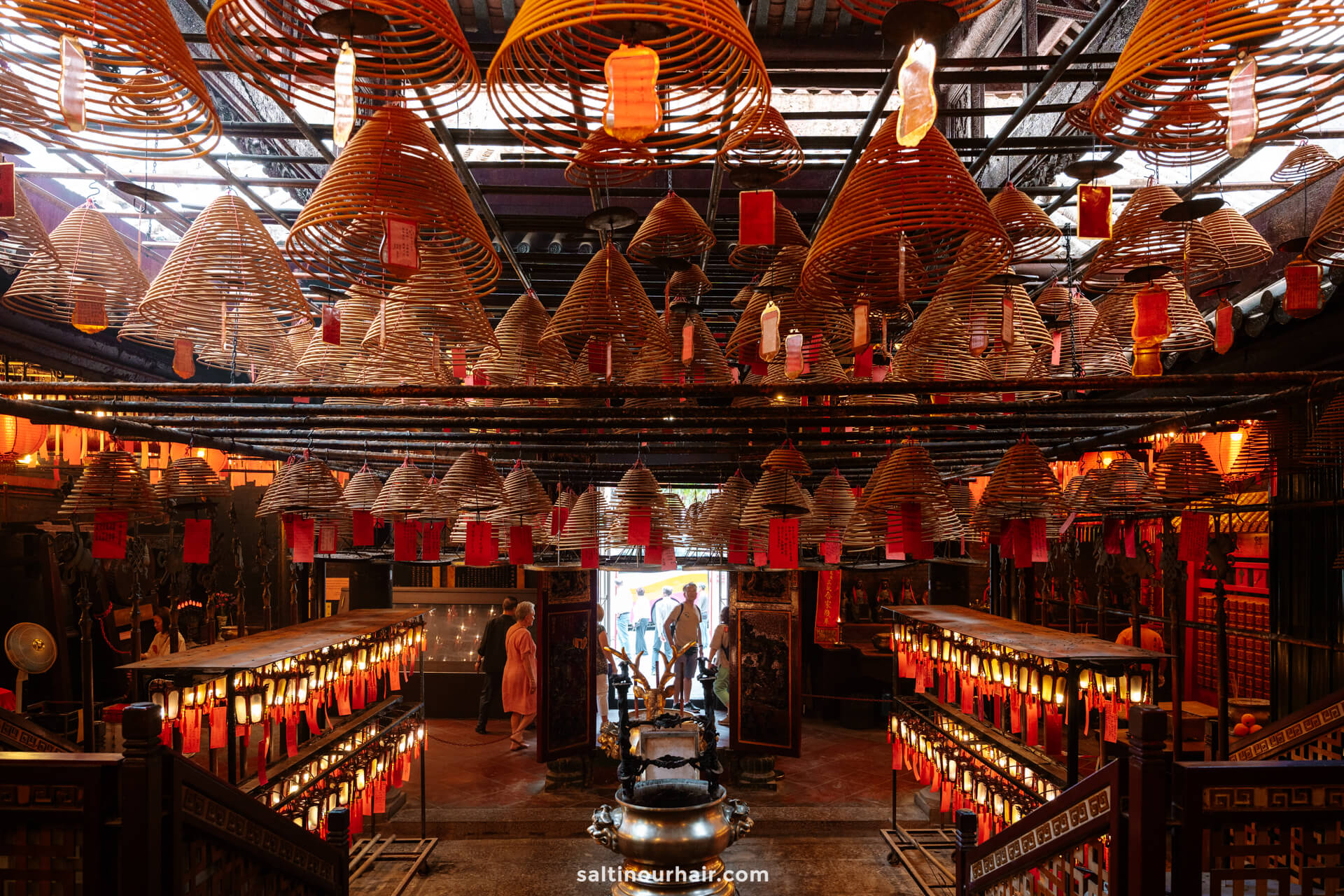 Interior of Man Mo in Hong Kong, with numerous hanging spiral incense coils, lanterns, and visitors entering through a doorway in the background.