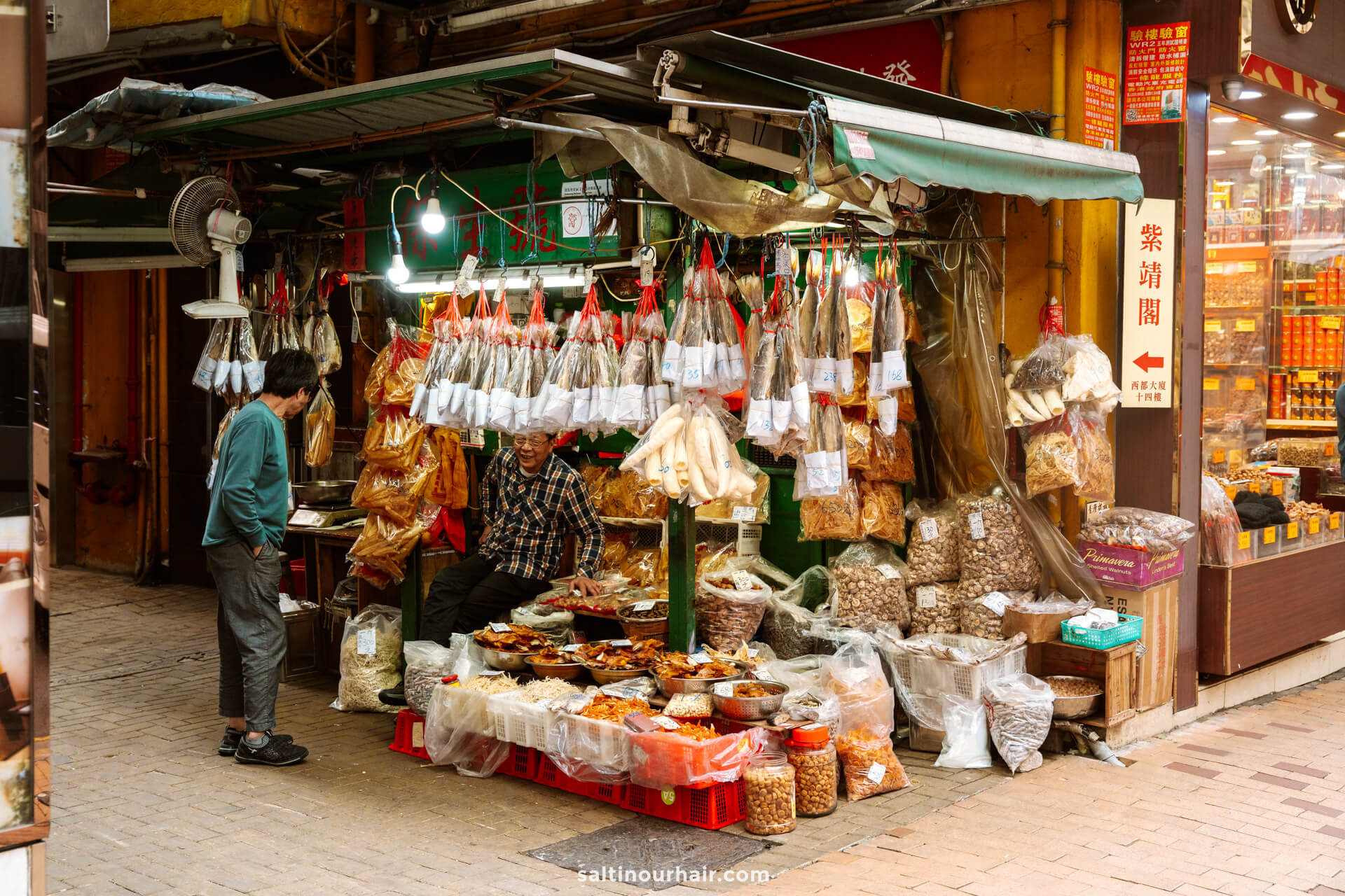 Two people converse at a bustling street market stall in Sheung Wan, Hong Kong selling dried goods and herbs, with clear plastic bags of products hanging above trays and jars on display.