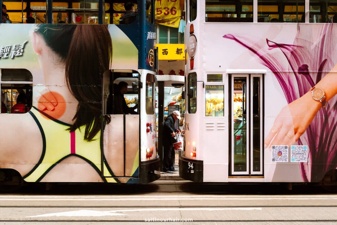 Two trams in Hong Kong with colorful advertisements stop side by side with pedestrians weaving between them.