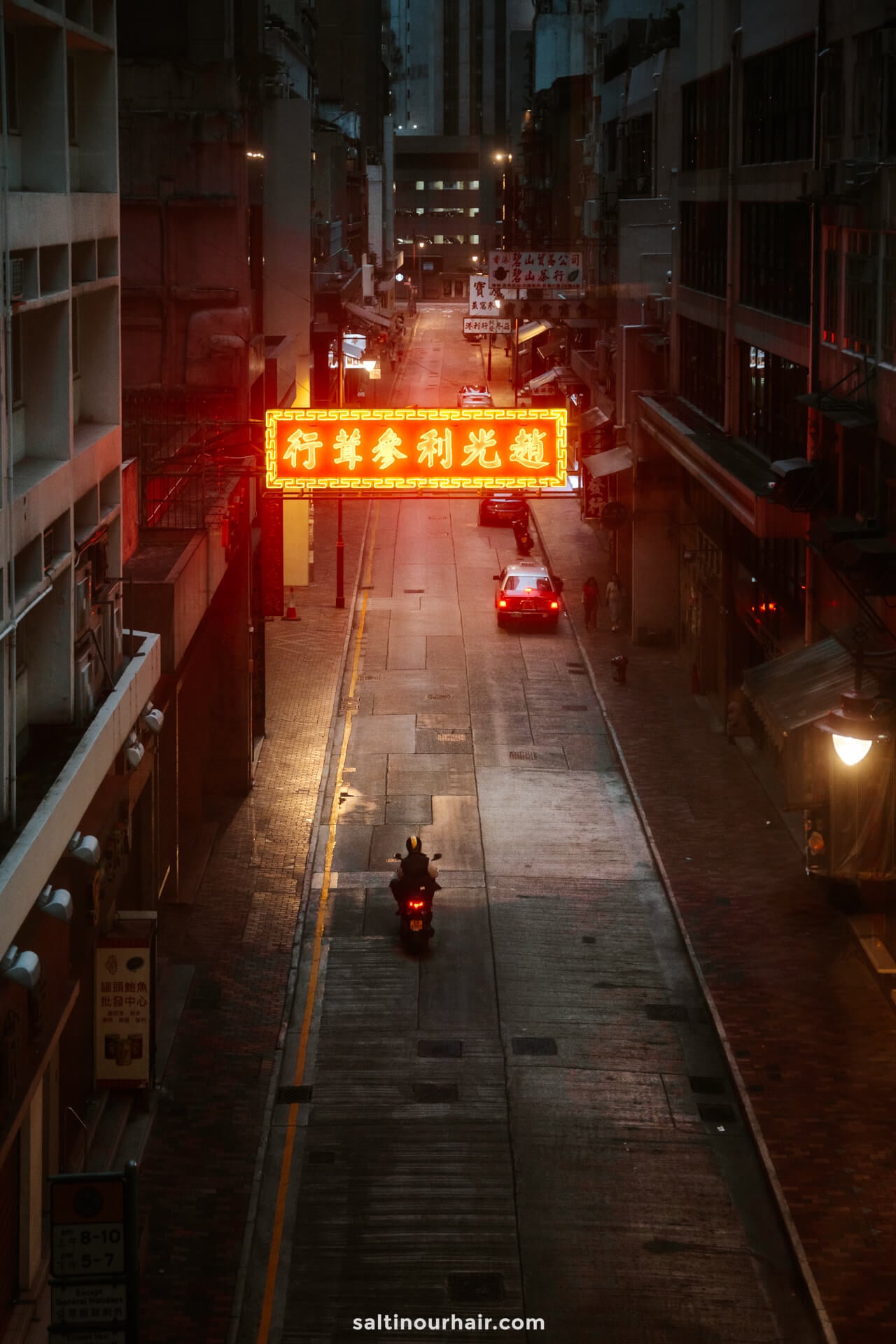A motorbike and a car travel down a dimly lit city street at dusk, with a glowing red neon sign featuring Chinese characters overhead