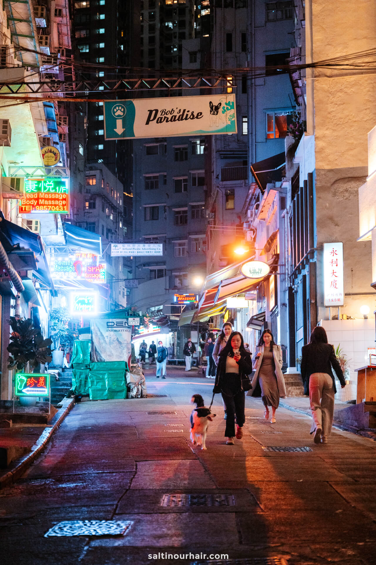 People walk along a brightly lit urban street at night, lined with neon signs and shopfronts&mdash;reminiscent of a vibrant hong kong itinerary.